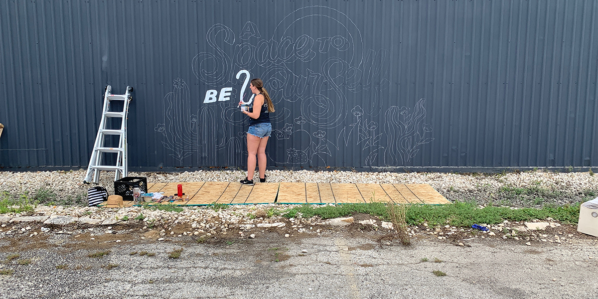 woman sets up to paint a mural on the side of a warehouse